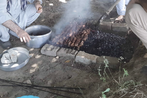 Photo of two people cooking meat over a charcoal stove. 