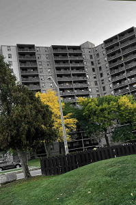 Black and white photo of a residential building. The only colour is green trees and grass. 